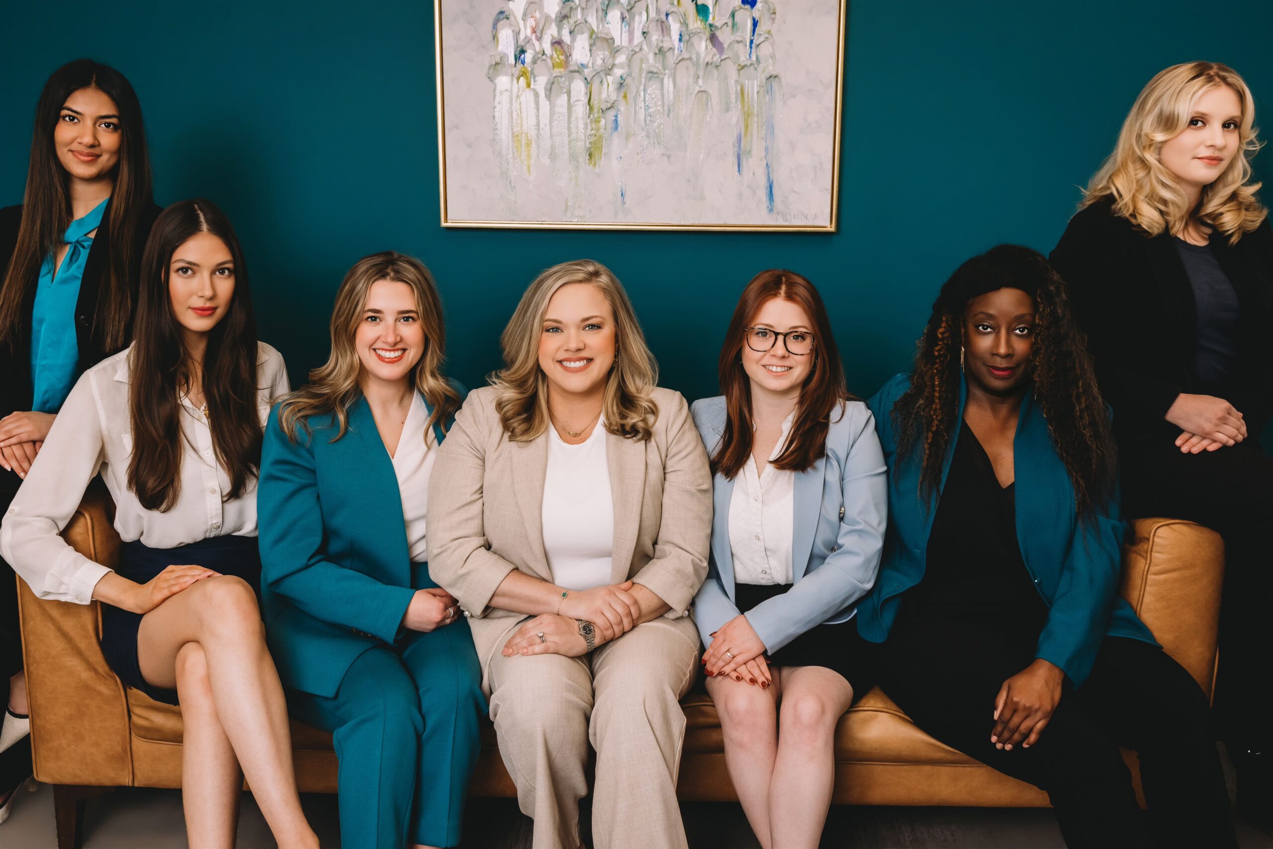 A group of seven professional women posing for a team portrait against a teal wall. Five women are seated on a tan leather sofa, and two are standing behind them.